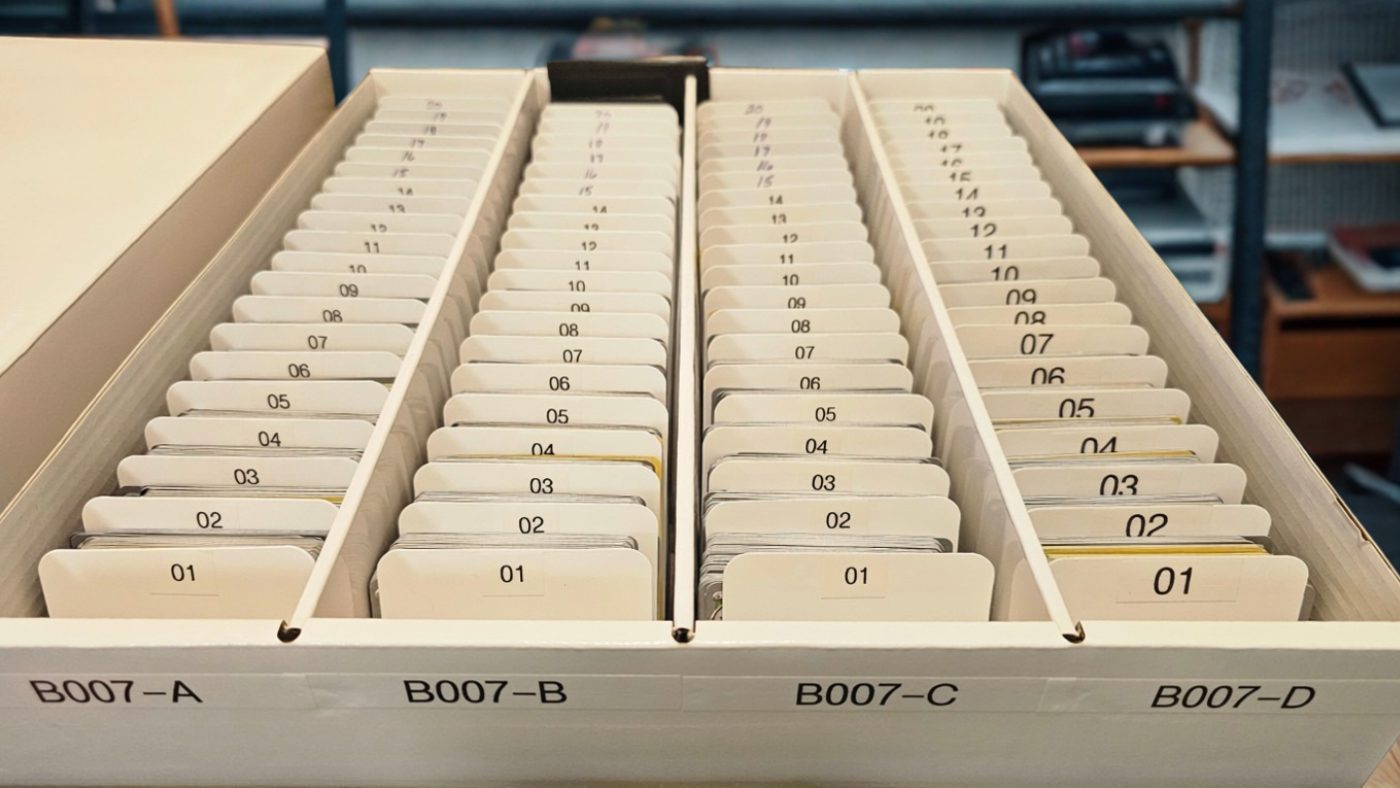 One storage box on a shelf, opened. Four rows of dividers run front
                  to back, each row labelled on the front edge of the box &mdash; B007-A,
                  B007-B, B007-C, B007-D &mdash; and each divider numbered 01 upward.
                  Items sit behind the dividers in no order but the one they arrived in.